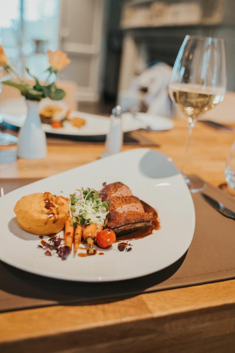 Gourmet-Erlebnis im Restaurant Giorgios im Hofgut Georgenthal: Hauptgericht mit Fleisch und Gemüse, Weinglas, Holztisch, elegante Atmosphäre mit Rosen und Kamin im Hintergrund.