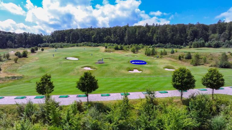 Malerischer Golfplatz Hofgut Georgenthal: Grüne Fairways, Sandbunker, Driving Range, umgeben von Wald, entspannte Atmosphäre unter blauem Himmel mit weißen Wolken.