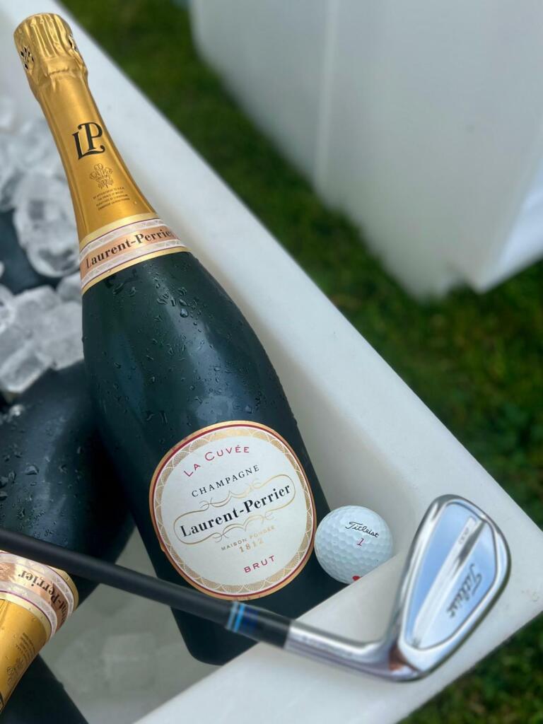 Champagner beim Hofgut Georgenthal: Gekühlte Laurent-Perrier Champagnerflasche in Eis, neben einem Titleist Golfball und einem Golf wedge, auf grünem Gras, entspannte Atmosphäre im Freien.