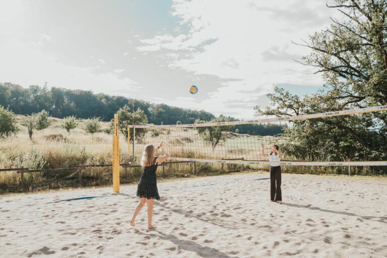 Zwei Personen spielen Beachvolleyball auf einem Sandplatz im Freien. Eine Person in einem schwarzen Kleid schlägt den Ball über das Netz, während die andere Person in einer weißen Bluse und dunklen Hosen auf der gegenüberliegenden Seite des Netzes wartet. Im Hintergrund erstreckt sich eine natürliche Landschaft mit Wiesen, Bäumen und einem bewölkten Himmel.