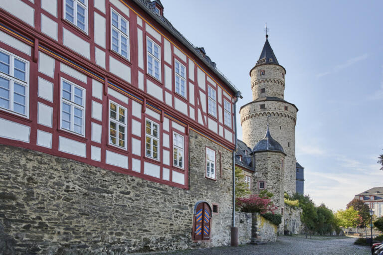 Ein Bild aus Idstein, Deutschland, das den Hexenturm und ein angrenzendes Fachwerkhaus zeigt. Der Hexenturm ist ein mittelalterlicher Wehrturm aus Stein mit einem spitzen Dach und kleinen Fenstern, der ein Wahrzeichen der Stadt ist. Daneben steht ein Fachwerkhaus mit weißem Putz und roten Holzbalken, typisch für die regionale Architektur.
