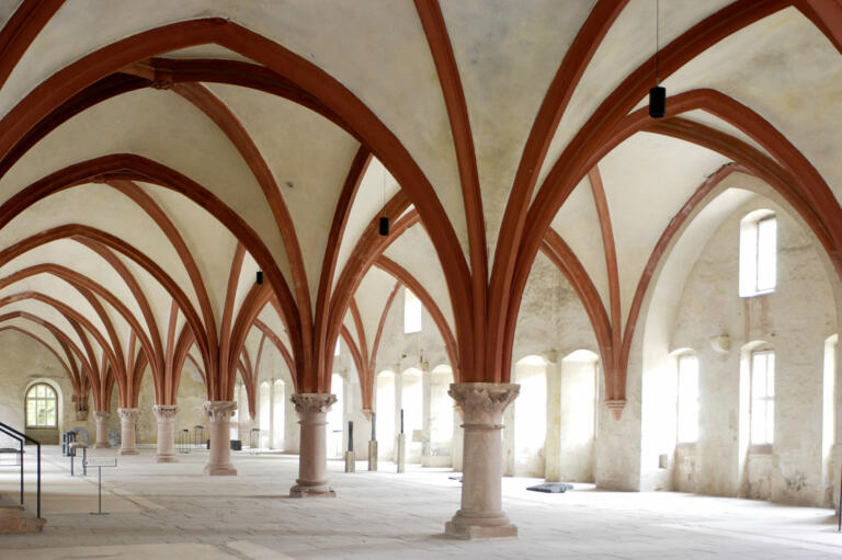 Ein Innenraum des Klosters Eberbach im Rheingau, Deutschland. Das Bild zeigt den Laienrefektoriumssaal mit seinen beeindruckenden gotischen Kreuzrippengewölben. Die hohen, gewölbten Decken werden von schlanken Säulen mit roten Holzbögen gestützt, die sich in regelmäßigen Abständen über den Raum erstrecken.