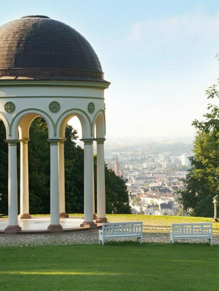 Ein Bild vom Neroberg in Wiesbaden, Deutschland. Im Vordergrund steht der Monopteros, ein kleiner, runder Pavillon mit einer dunklen Kuppel und weißen Säulen, der auf einer grünen Wiese thront.