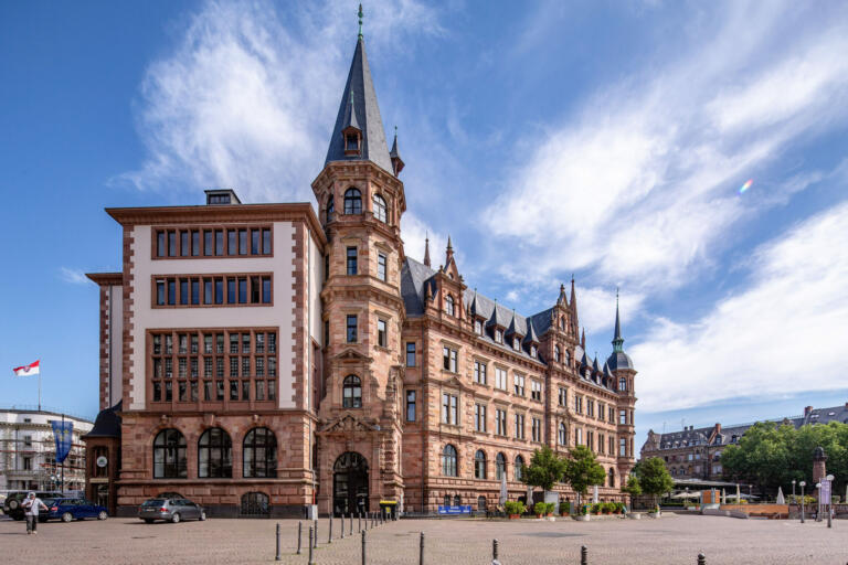 Ein Bild des Rathauses von Wiesbaden, Deutschland. Das Gebäude ist ein imposanter Bau im neugotischen Stil mit einer Fassade aus rotem Sandstein. Es hat mehrere Türme, von denen der höchste eine spitze Haube trägt, sowie zahlreiche Fenster mit gotischen Spitzbögen. Die Architektur ist detailreich mit Ziergiebeln und Ornamenten.