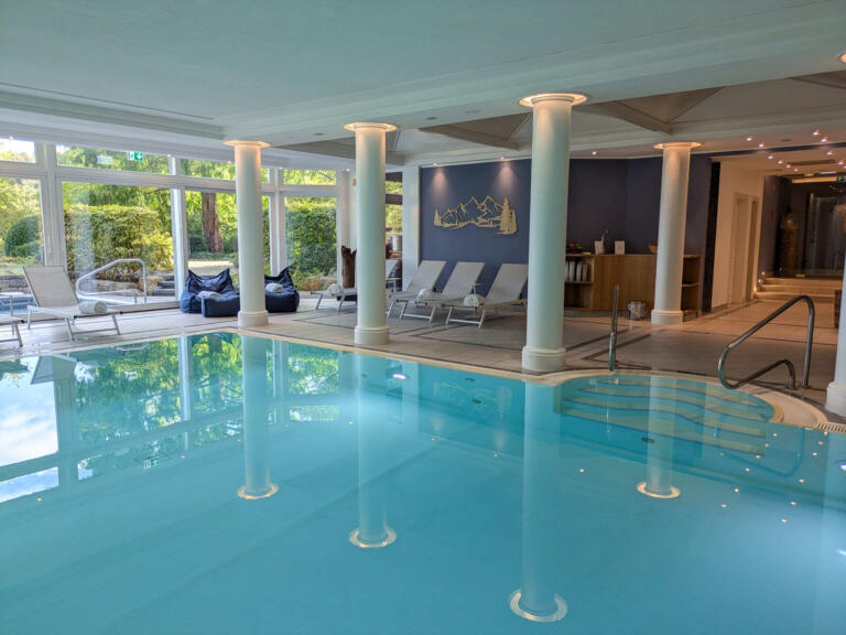 Der Wellnessbereich des Hofguts Georgenthal mit einem Innenpool, dessen türkisfarbenes Wasser sanft reflektiert. Um den Pool herum stehen mehrere Liegestühle, die zum Entspannen einladen. Große Fenster bieten einen Blick auf eine grüne Außenlandschaft mit Bäumen und Sträuchern.