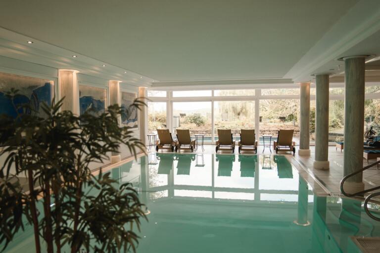 Der Wellnessbereich des Hofguts Georgenthal mit einem Innenpool, dessen türkisfarbenes Wasser sanft reflektiert. Um den Pool herum stehen mehrere gepolsterte Liegestühle, die zum Entspannen einladen. Große Fenster bieten einen Blick auf eine grüne Außenlandschaft mit Bäumen und Sträuchern.