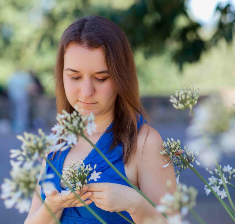 Eine Dame in einem blauen Kleid steht im Außenbereich des Hofguts Georgenthal und hält einen Strauß weißer Wildblumen in den Händen. Sie blickt auf die Blumen, während im Hintergrund unscharf eine grüne Landschaft mit Bäumen und Sträuchern zu sehen ist, was eine natürliche und friedliche Atmosphäre vermittelt.
