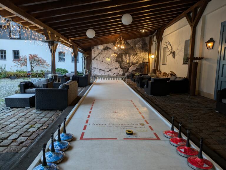 Ein überdachter Außenbereich mit einer Shuffleboard-Bahn in der Mitte. Die Bahn ist mit roten und blauen Markierungen versehen, und ein schwarzer Puck befindet sich darauf. Entlang der Bahn stehen rote und blaue Shuffleboard-Stöcke.