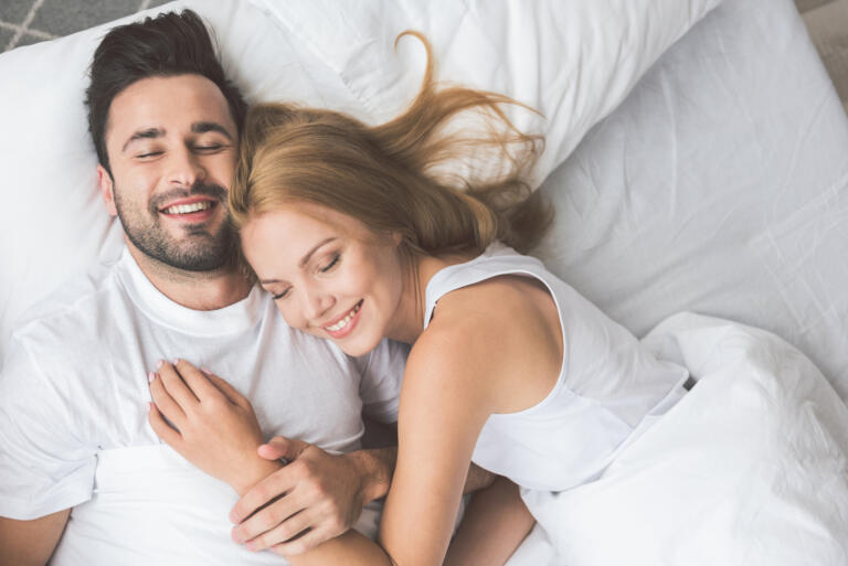 Entspannung im Hofgut Georgenthal: Ein Paar liegt eng umschlungen auf einem weißen Bett, umgeben von weichen Kissen und Decken, in einer ruhigen Atmosphäre.