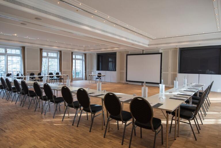 Konferenzraum im Hofgut Georgenthal mit einer U-förmigen Tischaufstellung, umgeben von schwarzen Stühlen. Große Fenster mit Vorhängen lassen Tageslicht herein, und ein großer Bildschirm hängt an der Wand gegenüber. Wasserflaschen und Notizblöcke stehen auf den Tischen.