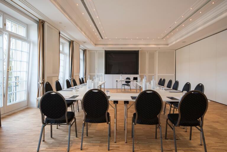 Konferenzraum im Hofgut Georgenthal mit einer U-förmigen Tischaufstellung, umgeben von schwarzen Stühlen. Große Fenster mit Vorhängen lassen Tageslicht herein, und ein großer Bildschirm hängt an der Wand gegenüber. Wasserflaschen und Notizblöcke stehen auf den Tischen.