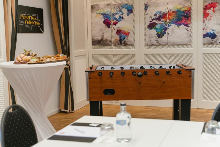 Besprechungsraum im Hofgut Georgenthal mit einem Kicker-Tisch, einem Stehtisch mit Snacks, einer Weltkarten-Wanddekoration und einem Schild mit der Aufschrift "Positive Thinking". Ein Tisch mit Tagungs-Setup, ausgestattet mit Wasserflaschen und Notizblöcken steht im Vordergrund.