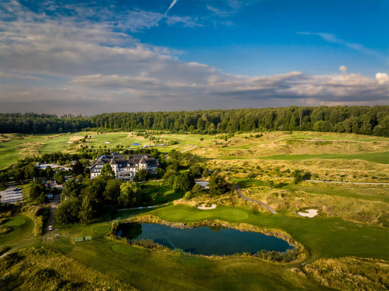 Luftaufnahme des Hofgut Georgenthal mit dem Hauptgebäude, umgeben von einem Golfplatz mit grünen Fairways, Sandbunkern, einem See und Wegen, eingebettet in eine waldreiche Landschaft unter einem bewölkten Himmel.