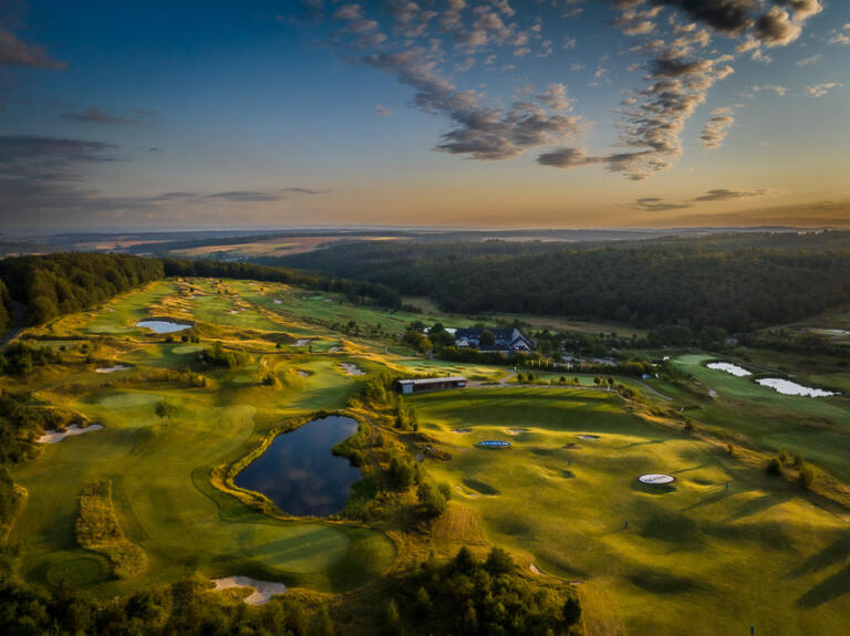 Luftaufnahme des Golfplatzes am Hofgut Georgenthal bei Sonnenuntergang, mit grünen Fairways, Sandbunkern, kleinen Seen und Wegen, umgeben von Wäldern und weiten Hügeln unter einem dramatischen Himmel.