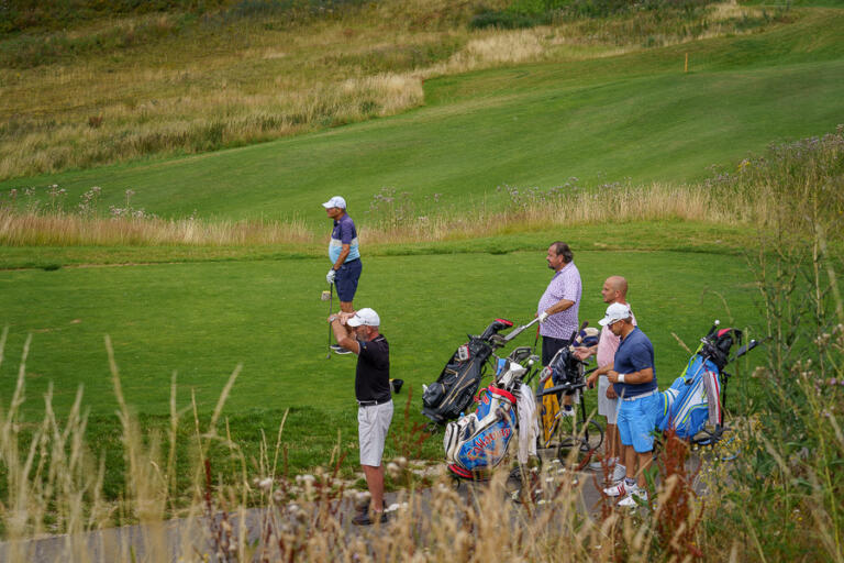 Gruppe von Golfern auf dem Golfplatz im Hofgut Georgenthal, einige mit Golftaschen, während einer Spieler schlägt. Grüne Fairways und Rough sind im Hintergrund sichtbar.