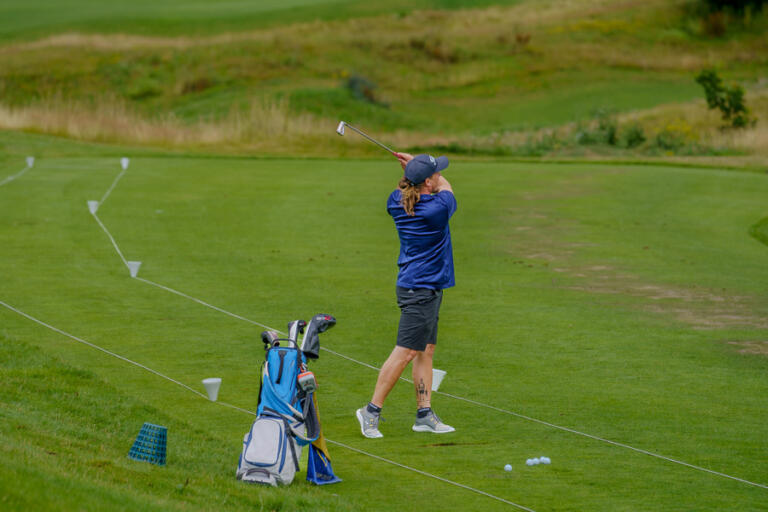Ein Golfspieler in blauer Kleidung schlägt auf der Driving Range des Hofgut Georgenthal ab, neben einer Golftasche und mehreren Bällen, umgeben von grünem Rasen und Hügeln.