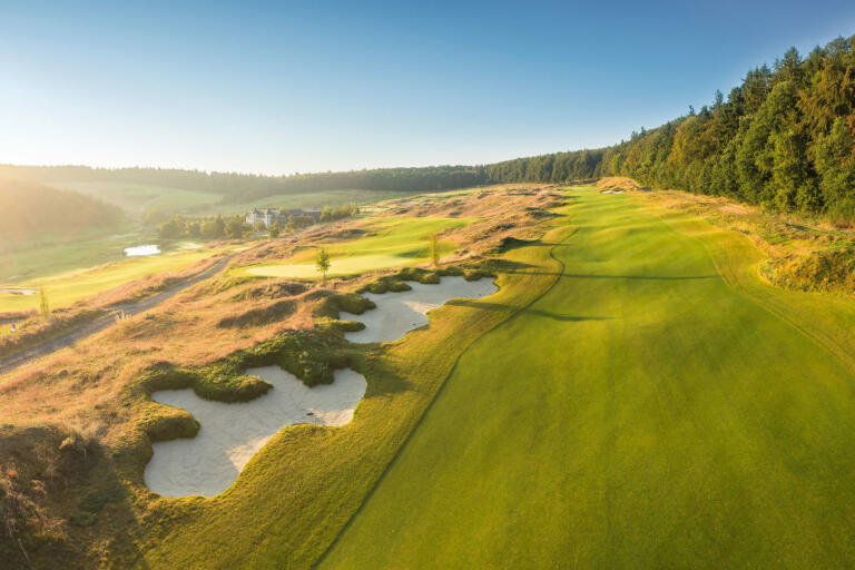 Golfplatz des Hofgut Georgenthal bei Sonnenschein: Grüne Fairways mit Sandbunkern, umgeben von Hügeln, Wäldern und einem kleinen Teich im Hintergrund.