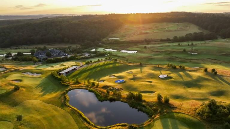 Luftaufnahme des Hofguts Georgenthal bei Sonnenuntergang, mit einem Golfplatz, einem herzförmigen Teich und umliegenden Wäldern.