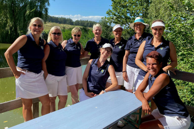 Die Damenmannschaft in blauen Golfshirts und weißen Shorts steht und sitzt an einem Holztisch am Hofgut Georgenthal, mit einem Teich und Bäumen im Hintergrund.