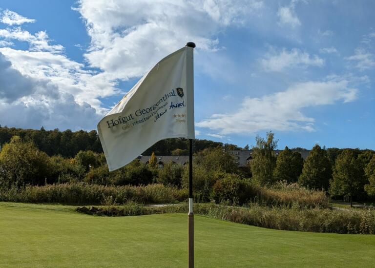 Flagge auf dem Hofgut Georgenthal: Weiße Fahne mit dem Logo 'Hofgut Georgenthal' weht auf einem Golfplatz, vor grünen Hügeln und Wäldern unter einem blauen Himmel.