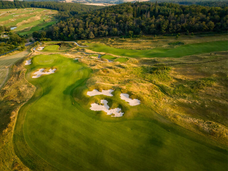 Luftaufnahme des Golfplatzes auf dem Hofgut Georgenthal: Grüne Fairways mit mehreren Sandbunkern, umgeben von Bäumen und Hügeln, bei warmem Sonnenlicht.