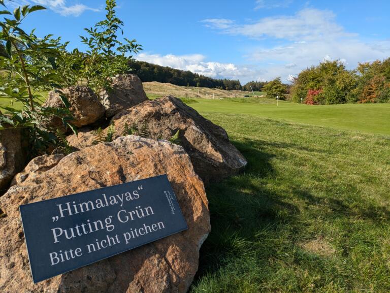 Ein Schild am Hofgut Georgenthal Golfplatz mit der Aufschrift 'Himalayas Grün – Putting pitchen bitte nicht', befestigt an einem großen Felsen, vor einem grünen Fairway mit Hügeln und Wäldern im Hintergrund.
