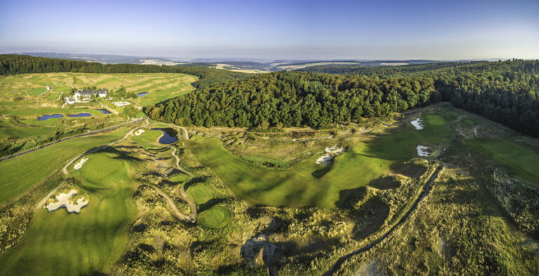 Panorama-Luftaufnahme des Golfplatzes am Hofgut Georgenthal mit grünen Fairways, Sandbunkern, Wegen, kleinen Seen und einem Gebäude, umgeben von dichten Wäldern und weiten Hügeln unter einem klaren Himmel.