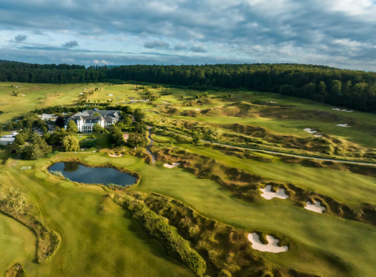 Luftaufnahme des Hofguts Georgenthal mit dem Hotelgebäude, einem Golfplatz, einem Teich und umliegenden Wäldern bei sonnigem Wetter.