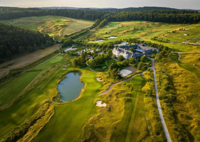Luftaufnahme des Hofguts Georgenthal mit dem Hotelgebäude, einem Golfplatz, einem Teich und umliegenden Wäldern bei sonnigem Wetter.