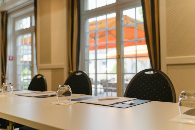 Konferenzraum im Hofgut Georgenthal mit Stühlen, Gläsern und Notizblöcken auf einem Tisch, großen Fenstern und Blick auf eine Terrasse mit orangefarbenem Sonnenschirm.