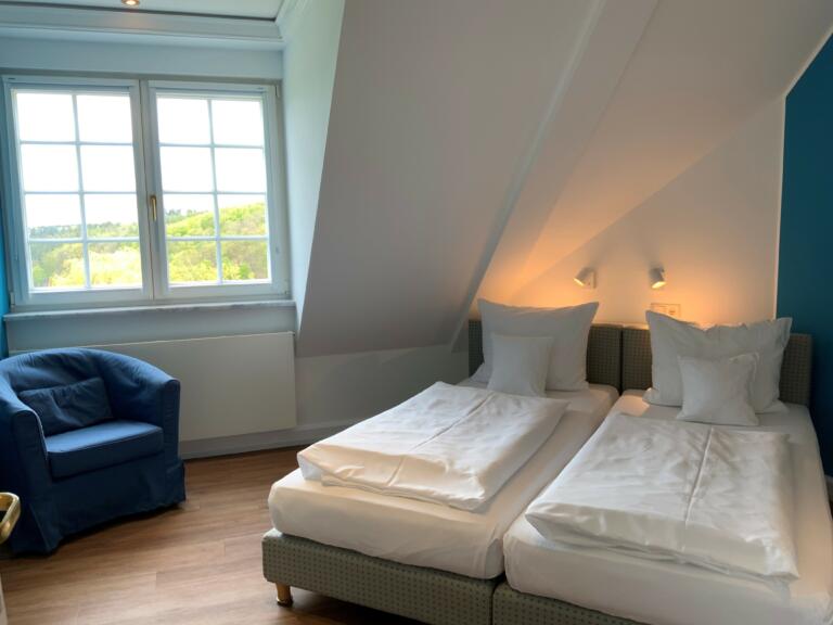 Hotelzimmer im Hofgut Georgenthal mit zwei Betten, einem blauen Sessel, Holzboden und einem großen Fenster mit Blick auf die Landschaft.