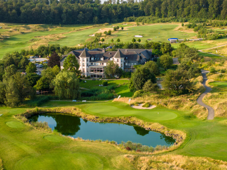 Luftaufnahme des Hofguts Georgenthal mit dem Hotelgebäude, einem Golfplatz, einem Teich und umliegenden Wäldern bei sonnigem Wetter.